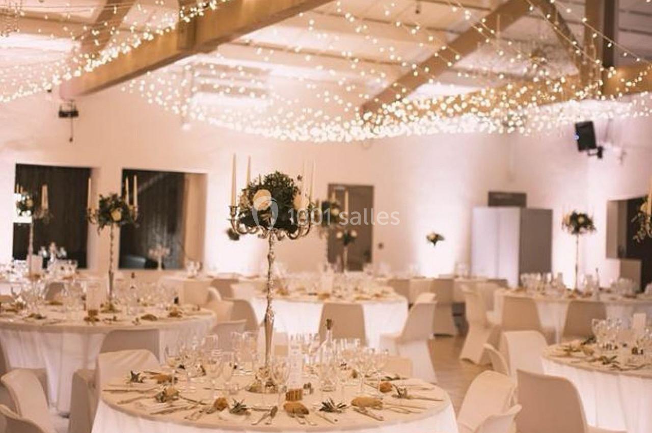 Salle de réception décorée pour un événement, avec tables rondes dressées, chandeliers et guirlandes lumineuses suspendues.