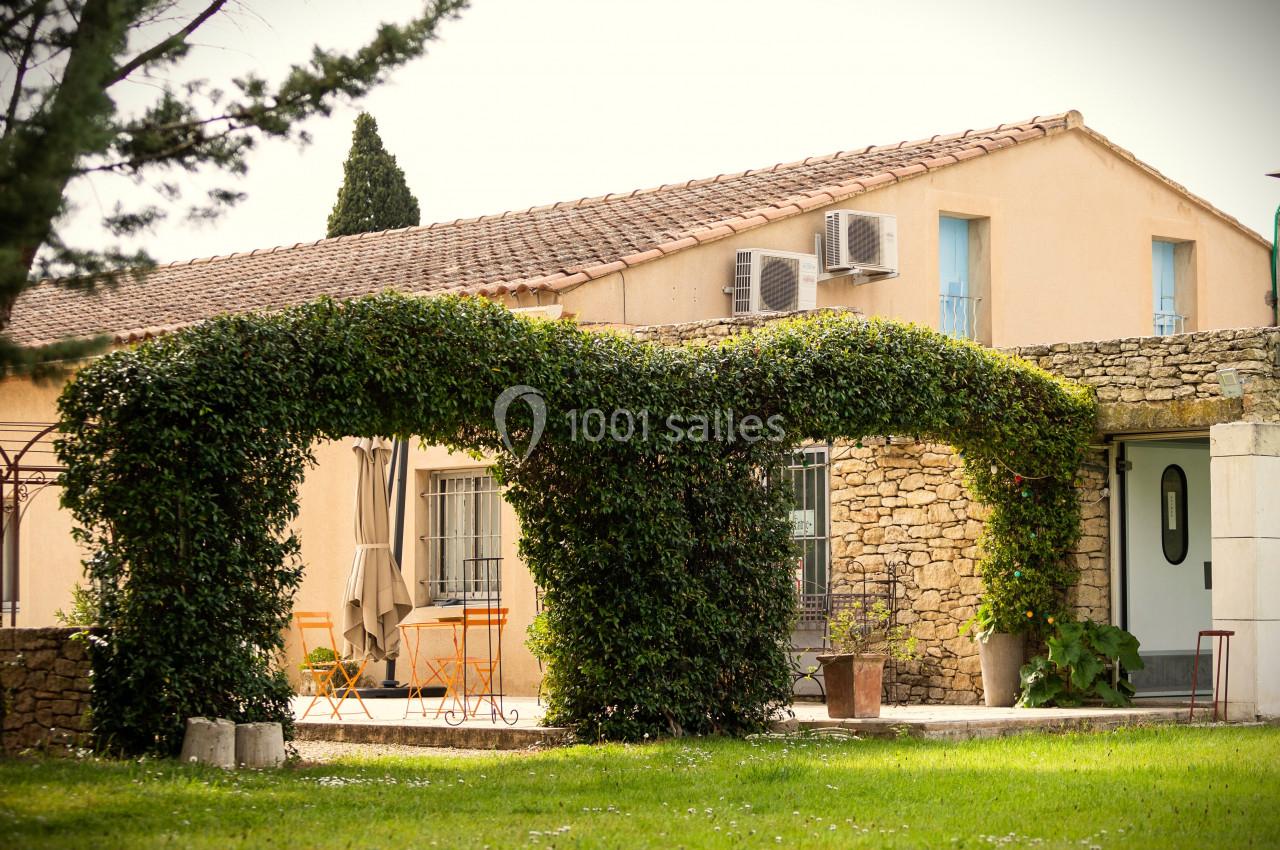 Arche végétale en lierre devant une maison en pierre avec terrasse aménagée et mobilier de jardin.