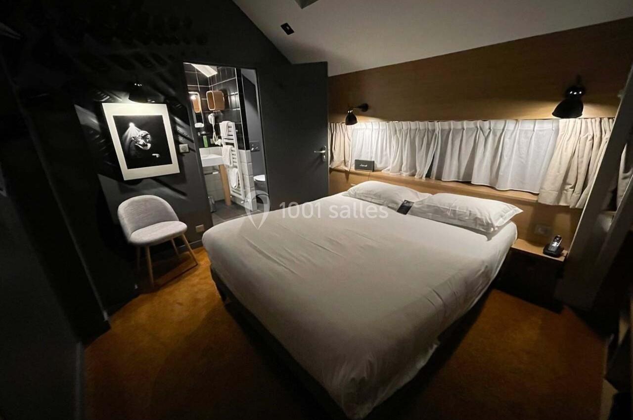 Chambre moderne avec lit double, éclairage tamisé, chaise, tableau au mur et salle de bain visible en arrière-plan.