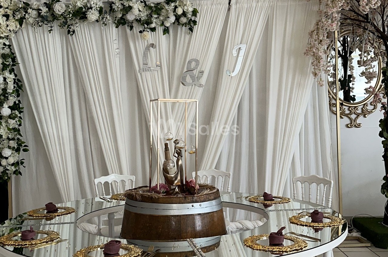 Table décorée pour un événement, avec nappes blanches, chaises assorties, centre de table floral et assiettes dorées.
