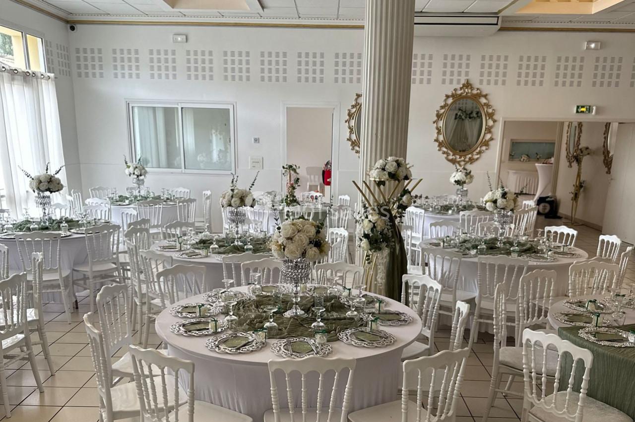 Salle de réception élégante avec tables rondes dressées, décorations florales blanches et chaises blanches.