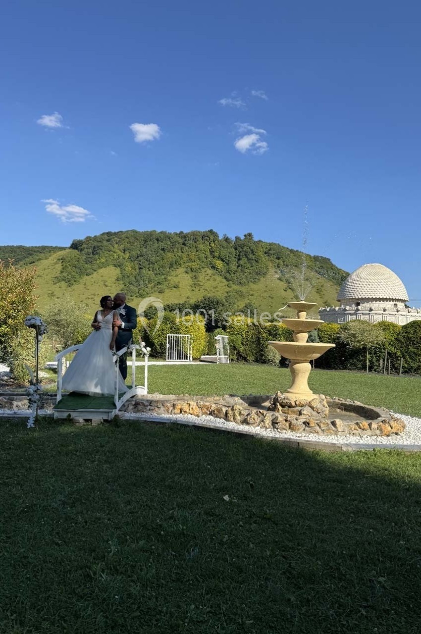 Un couple en tenue de mariage marche dans un jardin verdoyant avec une fontaine et une colline en arrière-plan.