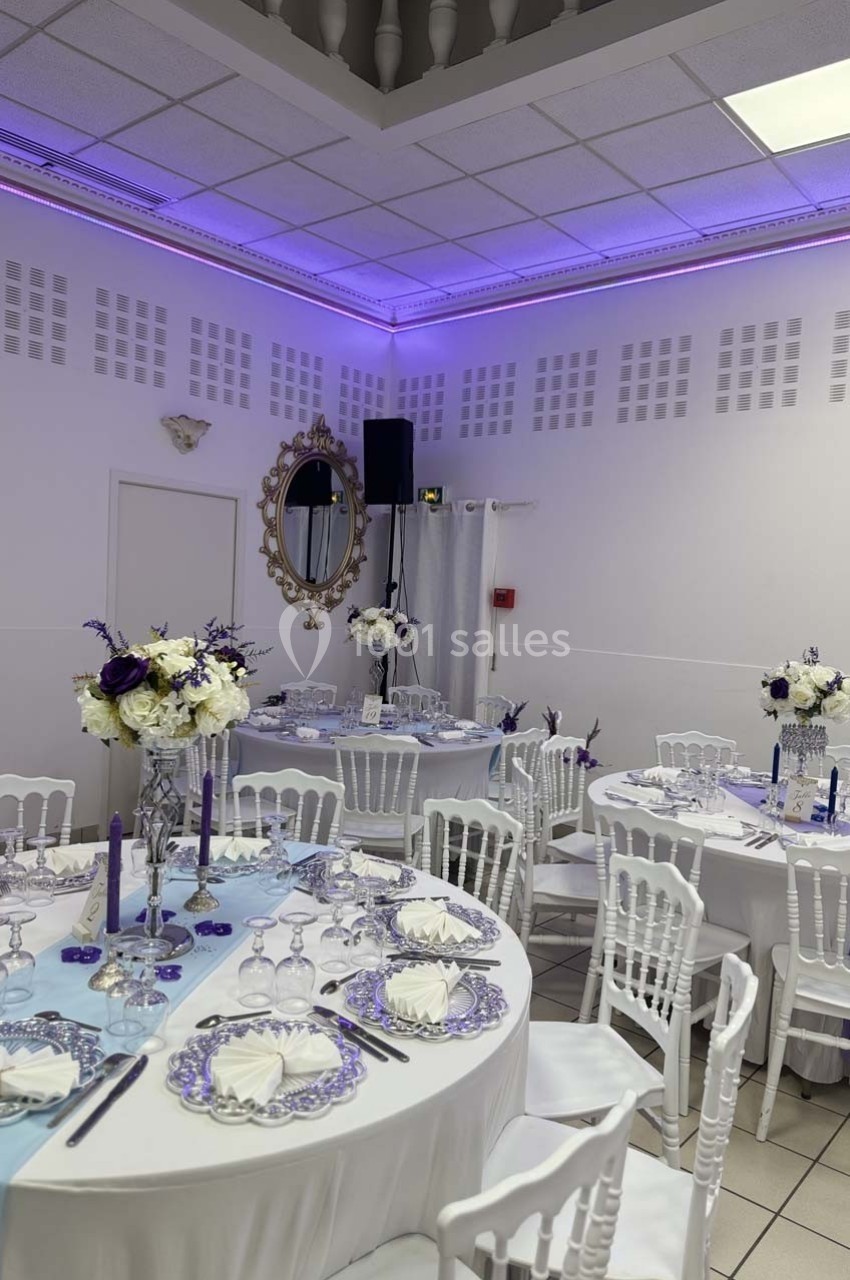 Salle de réception décorée avec des tables rondes, nappes blanches, centres de table fleuris et éclairage violet.