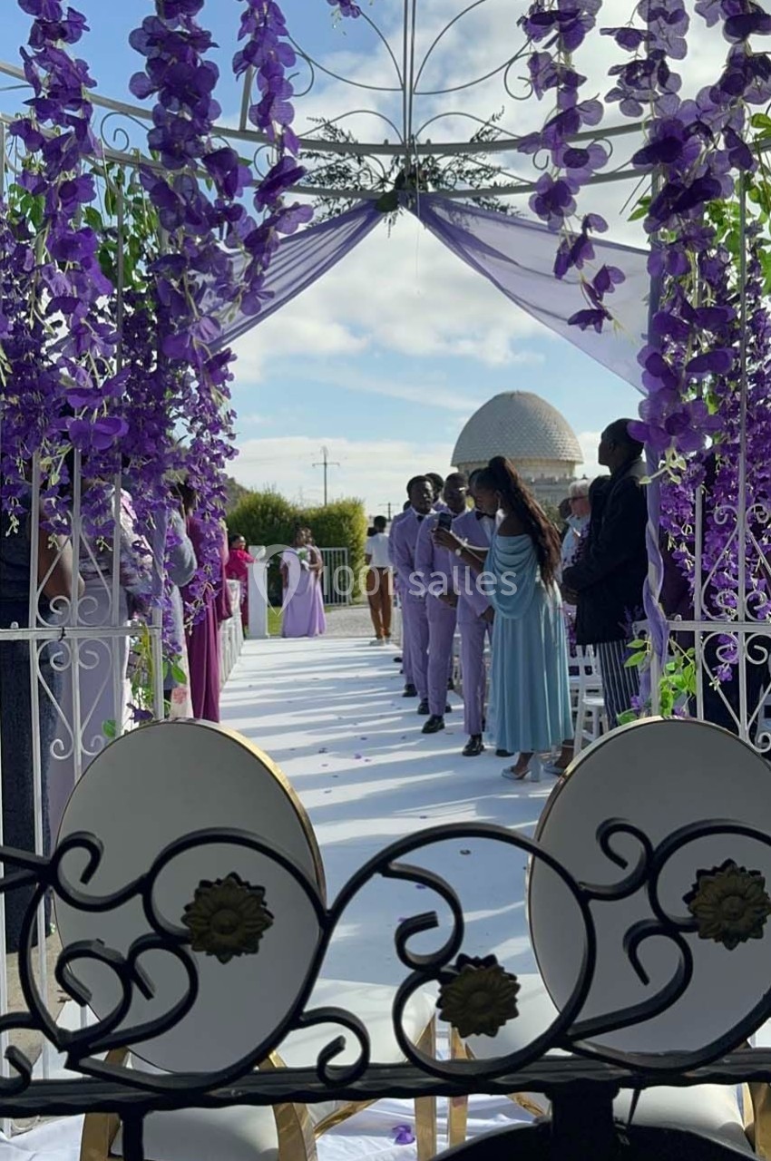 Allée décorée de fleurs violettes menant à une cérémonie en extérieur sous une arche, avec des invités en tenue élégante.
