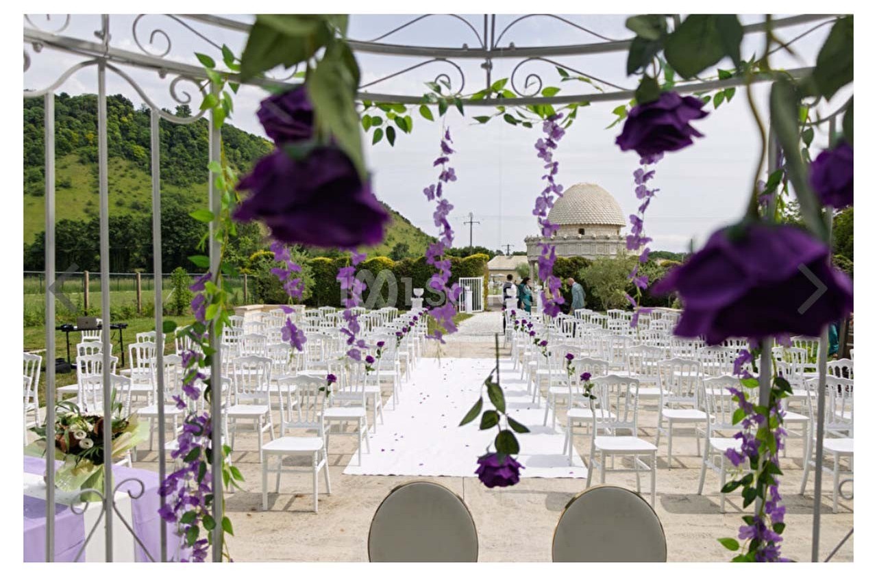Allée centrale décorée de fleurs violettes menant à un espace de cérémonie en plein air avec des chaises blanches alignées.