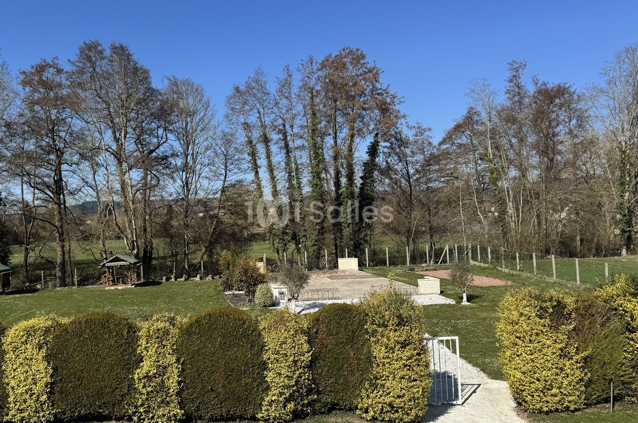Vue d'un jardin arboré avec pelouse, haies taillées, allée en gravier et arbres en arrière-plan sous un ciel bleu.