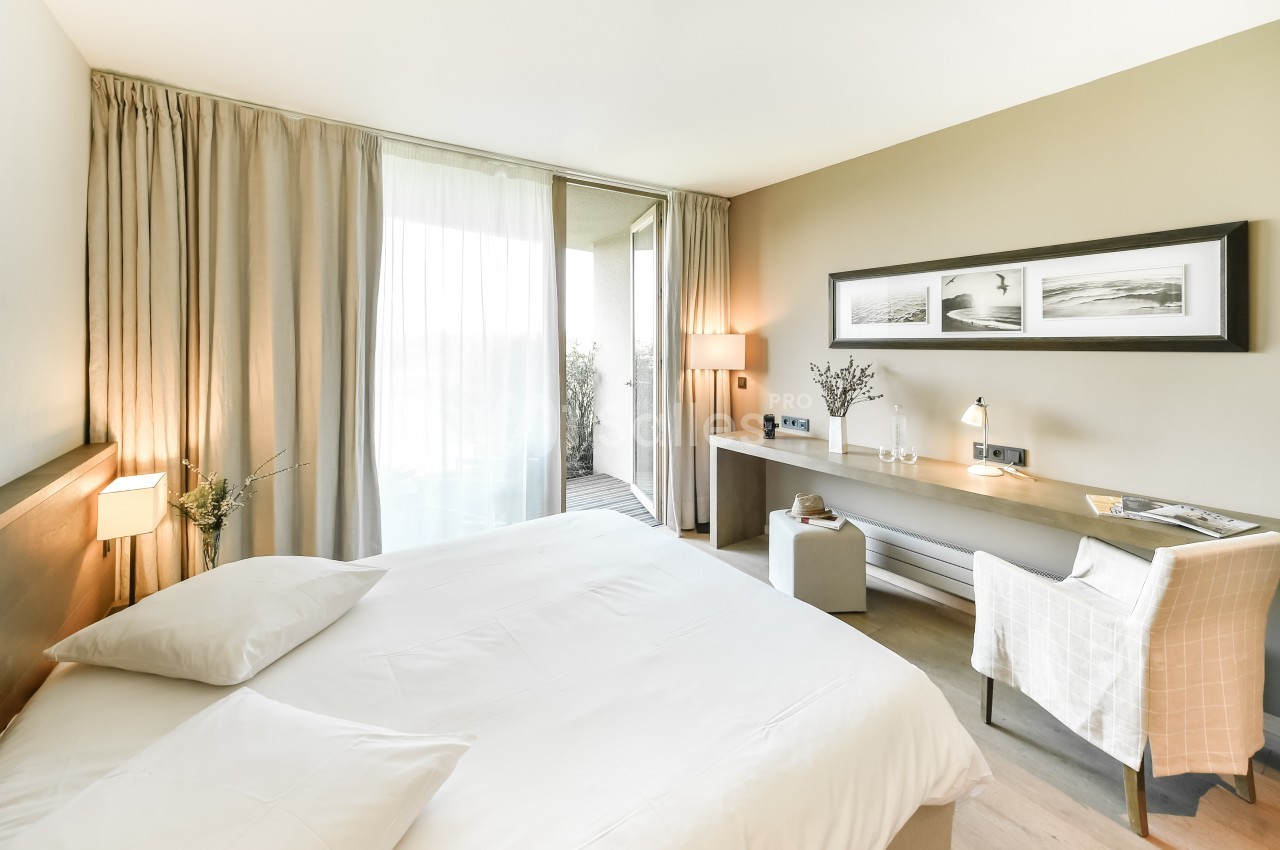 Chambre lumineuse avec lit double, bureau, chaise, rideaux beiges et porte-fenêtre donnant sur un balcon.