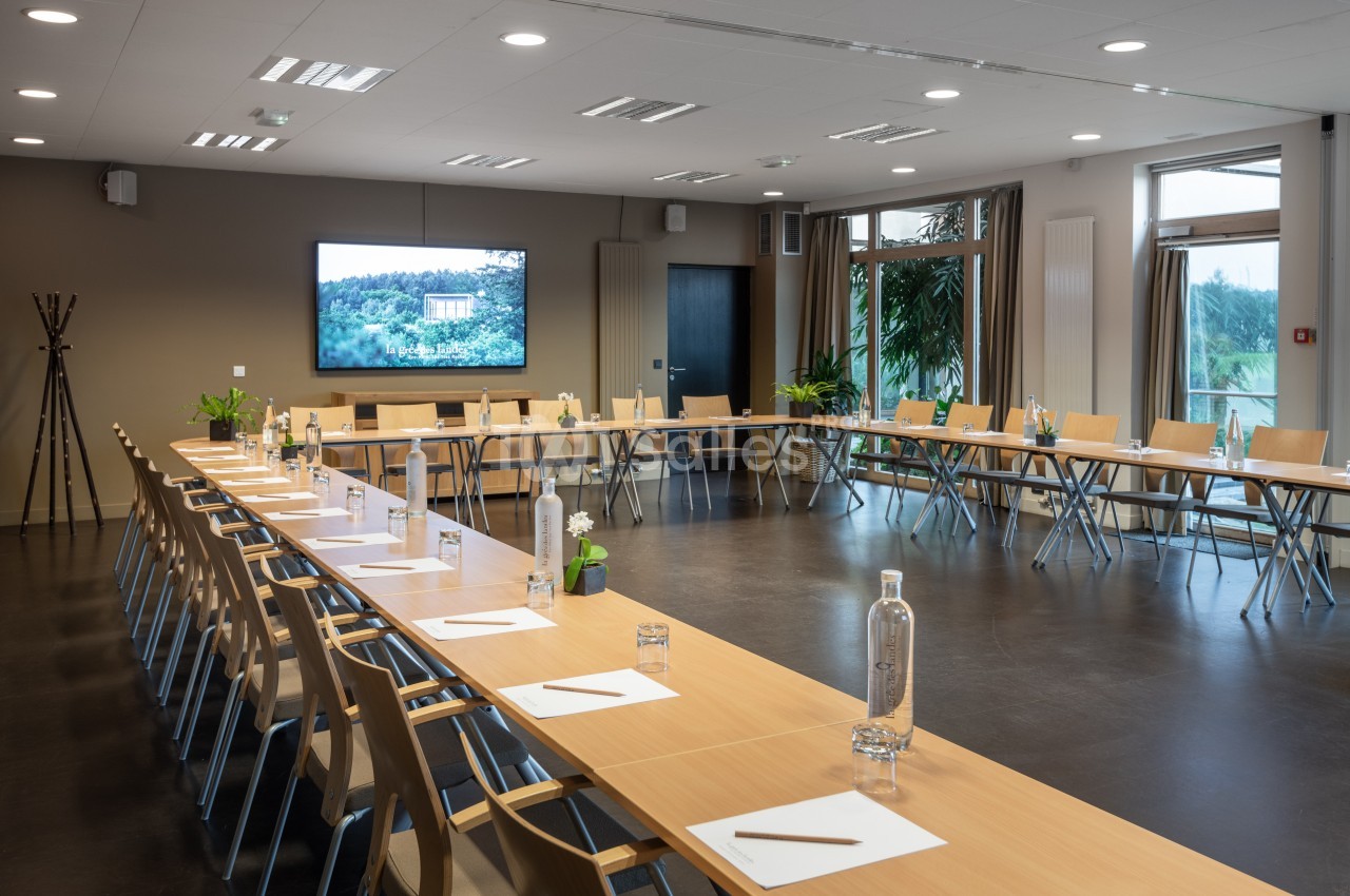 Salle de réunion lumineuse avec tables en U, chaises en bois, bouteilles d'eau, blocs-notes et écran affichant un paysage.