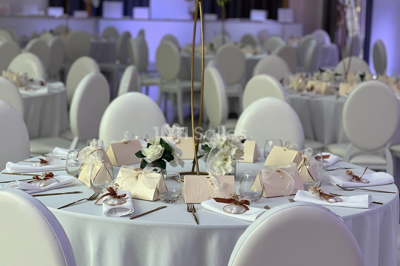 Table ronde élégamment dressée avec nappes blanches, décorations florales et cadeaux d'invités dans une salle de réception.