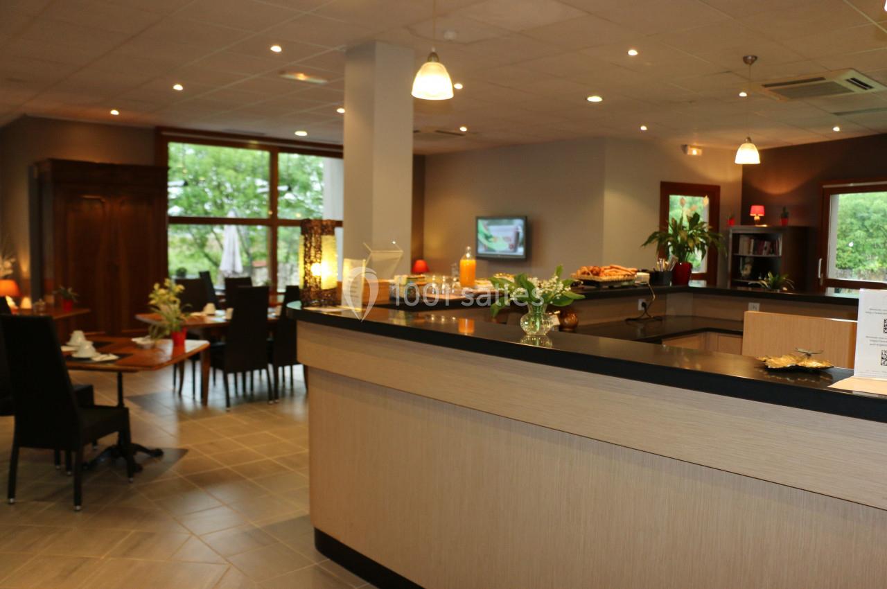 Salle de restaurant lumineuse avec comptoir en bois, tables dressées et grandes fenêtres donnant sur un espace vert.