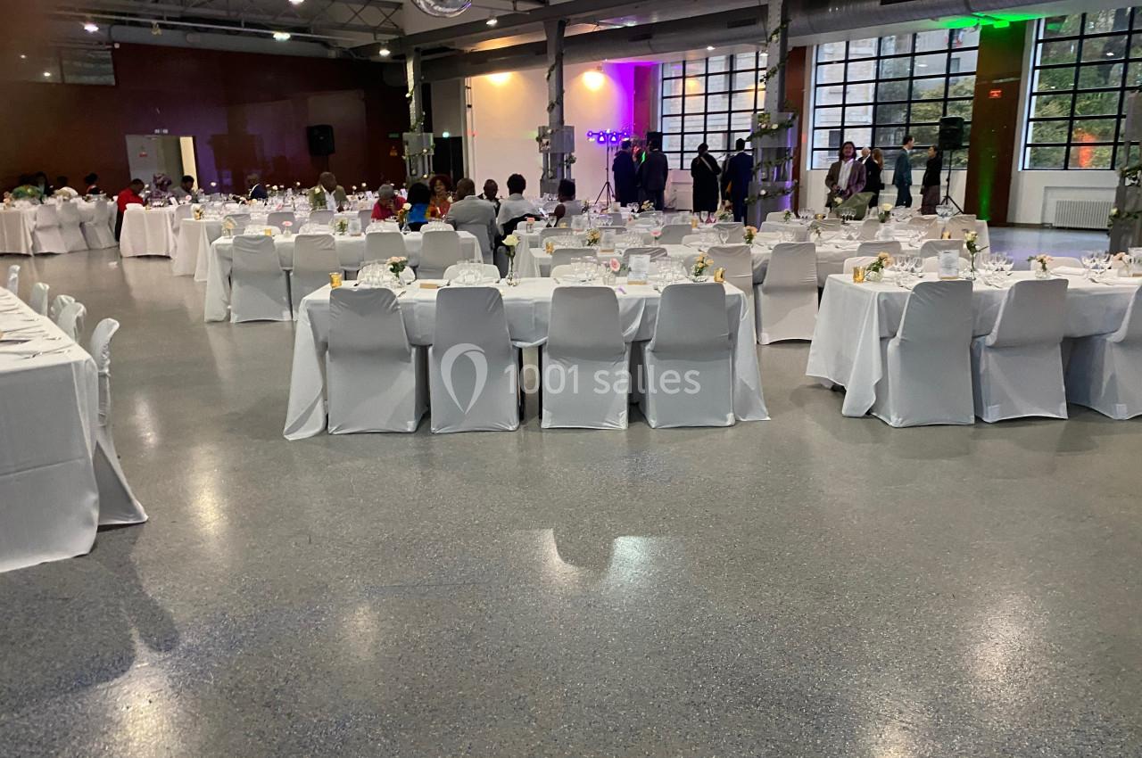 Salle de réception avec des tables rondes et rectangulaires dressées, chaises blanches et invités en arrière-plan.