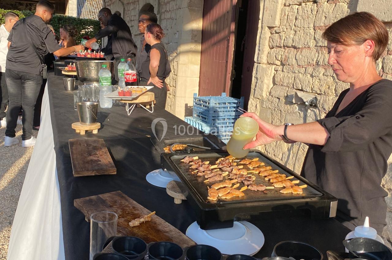 Une femme cuisine des brochettes sur une plancha lors d'un buffet en extérieur, avec des boissons et des plats disposés sur…