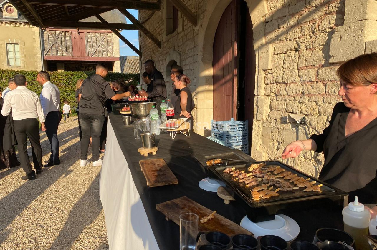Buffet en extérieur avec des plats chauds préparés sur une plancha, invités discutant près d'un bâtiment en pierre.