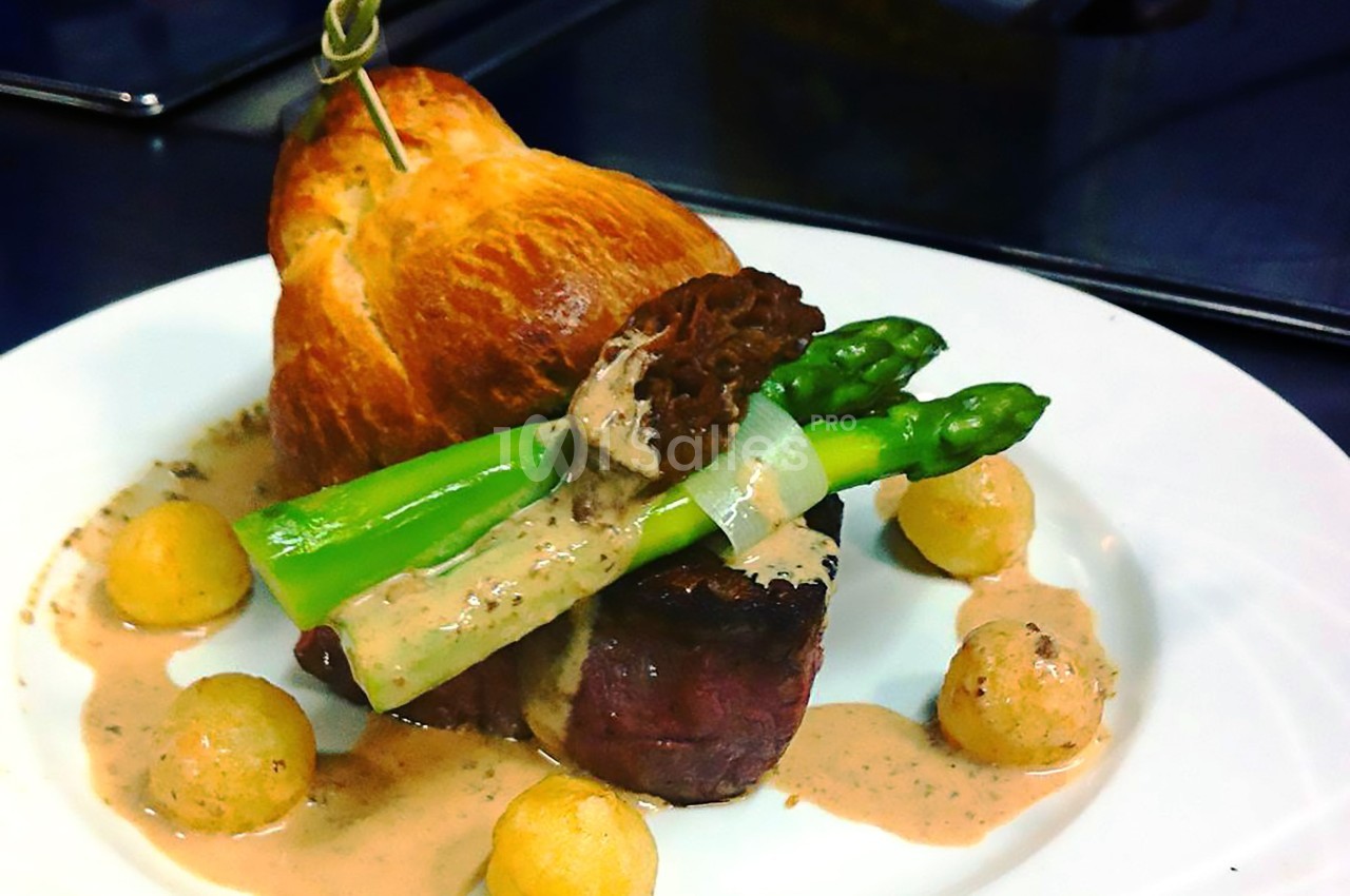 Assiette gastronomique avec filet de viande, asperges, morille, sauce crémeuse, pommes dauphines et brioche en arrière-plan.