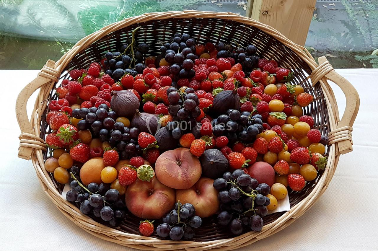 Panier en osier rempli de fruits variés, dont raisins, pêches, figues, fraises et prunes, posé sur une nappe blanche.