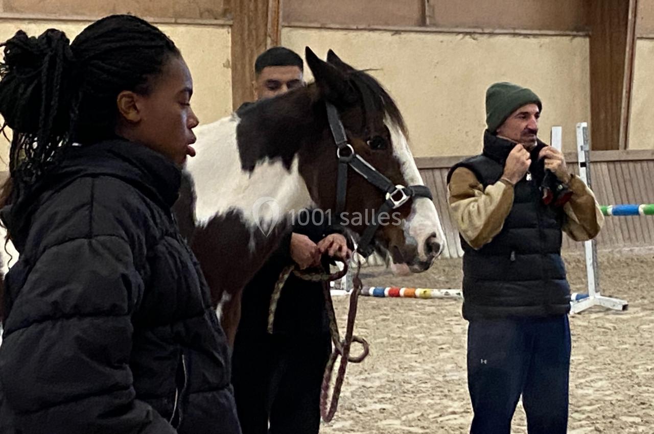 Deux personnes encadrent un cheval dans un manège équestre, une troisième ajuste son bonnet à l'arrière-plan.