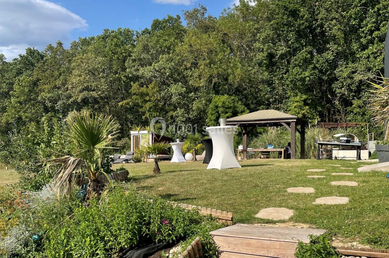 Jardin avec pelouse, terrasse en bois, tonnelle, tables hautes et végétation dense en arrière-plan.