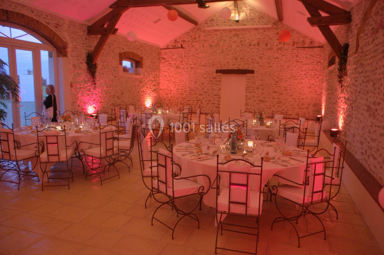 Salle en pierre avec des tables rondes dressées pour un événement, éclairée par des lumières tamisées roses.
