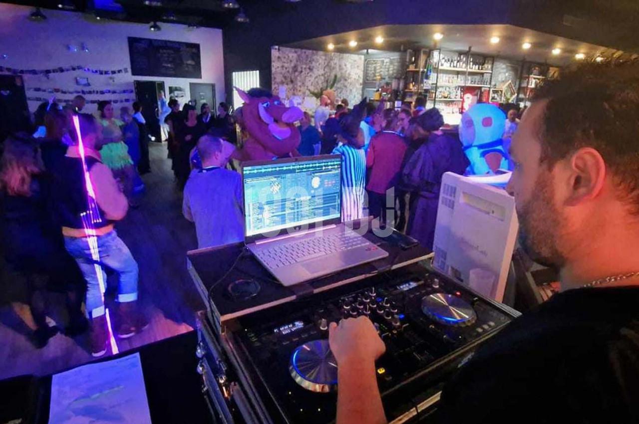 Un DJ mixe de la musique dans un bar animé avec des lumières colorées et des personnes dansant en arrière-plan.