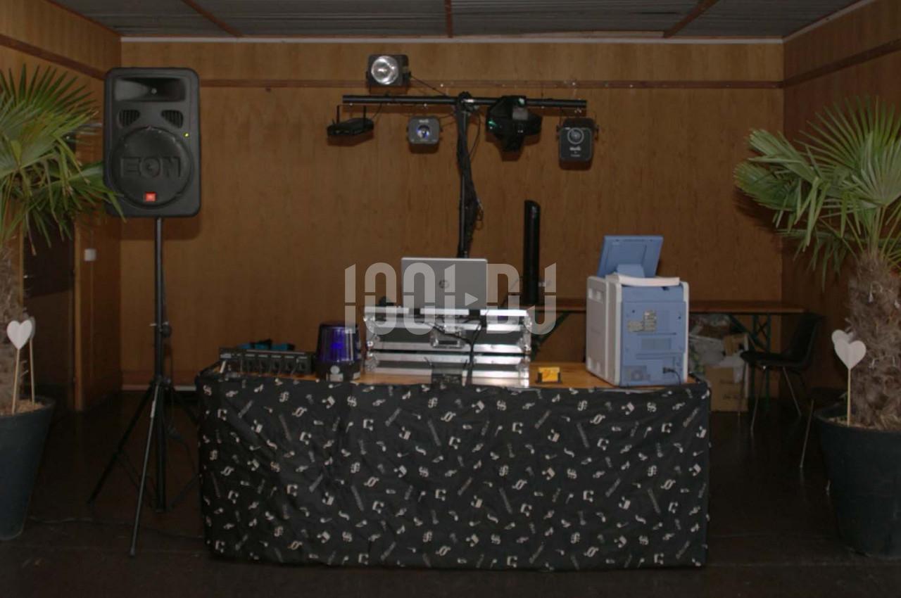 Table de mixage avec équipement audio et éclairage, encadrée par deux plantes en pot, dans une salle au mur en bois.