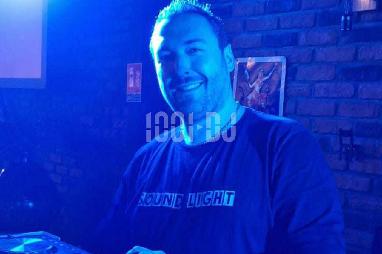 Homme souriant dans une ambiance lumineuse bleutée, debout devant un mur de briques avec un équipement audio visible.