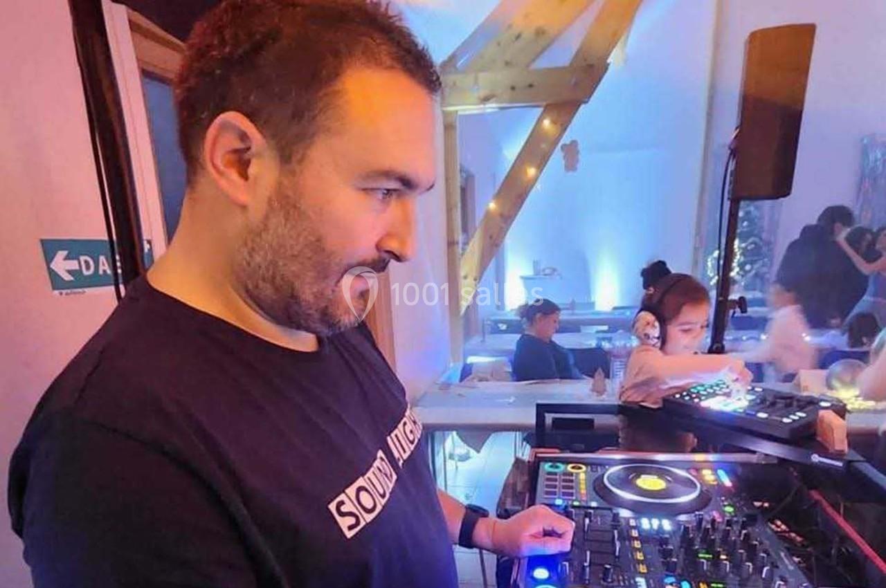 Un DJ mixe de la musique sur une console dans une salle éclairée, avec des personnes en arrière-plan.