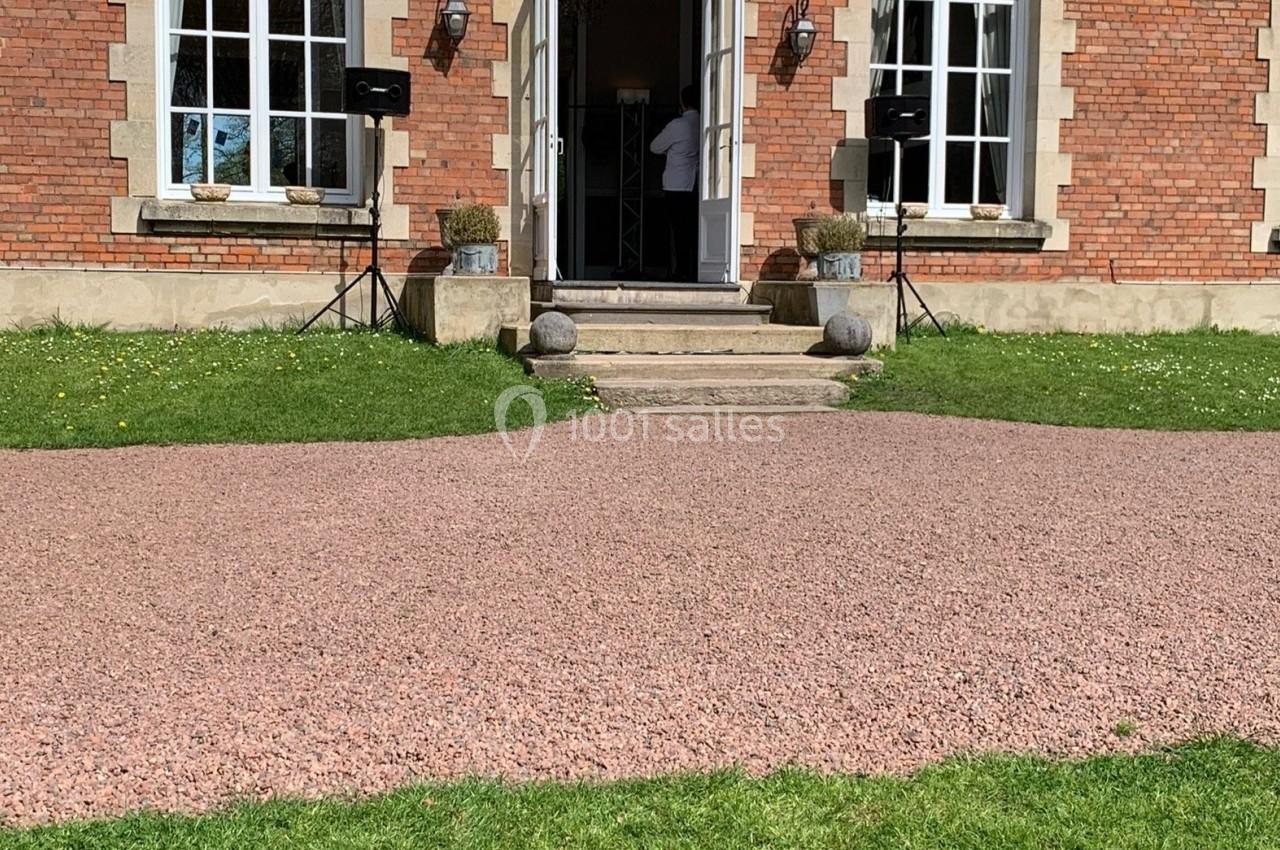 Entrée d'un bâtiment en briques avec porte ouverte, escaliers en pierre, pelouse et allée en gravier au premier plan.
