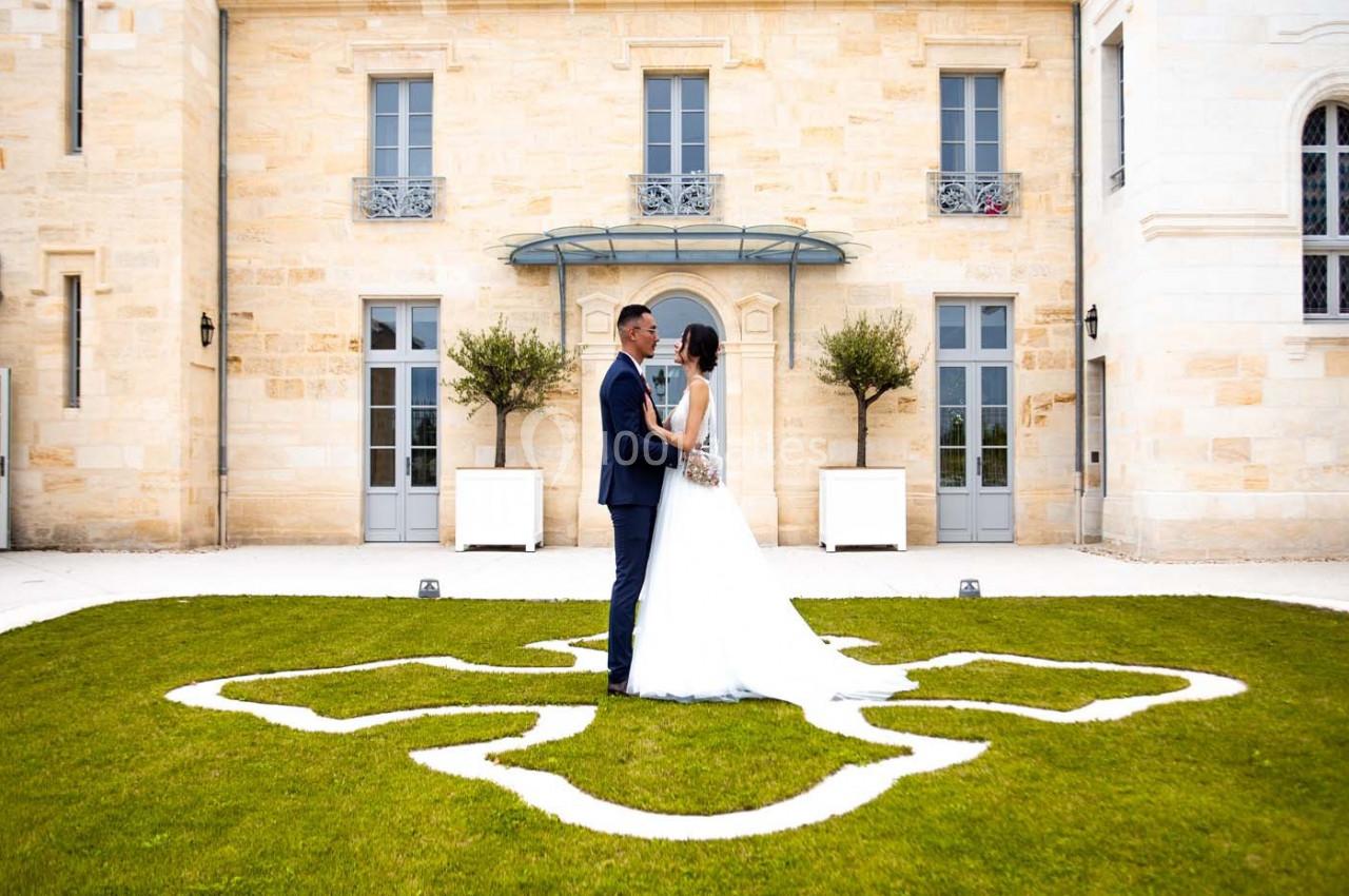 Un couple en tenue de mariage se tient face à face sur une pelouse ornée d'un motif blanc devant un bâtiment en pierre.