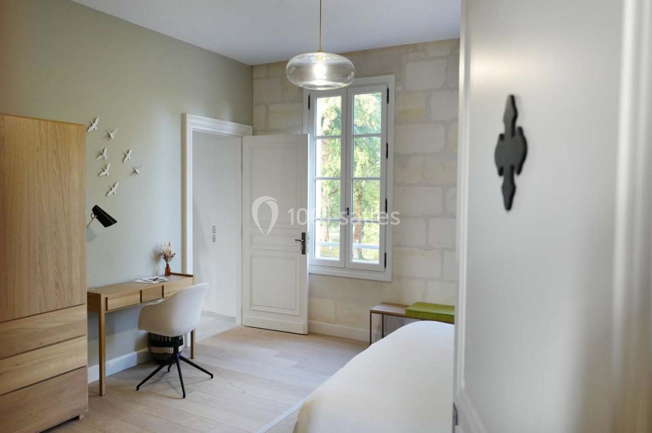 Chambre lumineuse avec bureau en bois, chaise blanche, armoire, lit et fenêtre donnant sur un extérieur verdoyant.