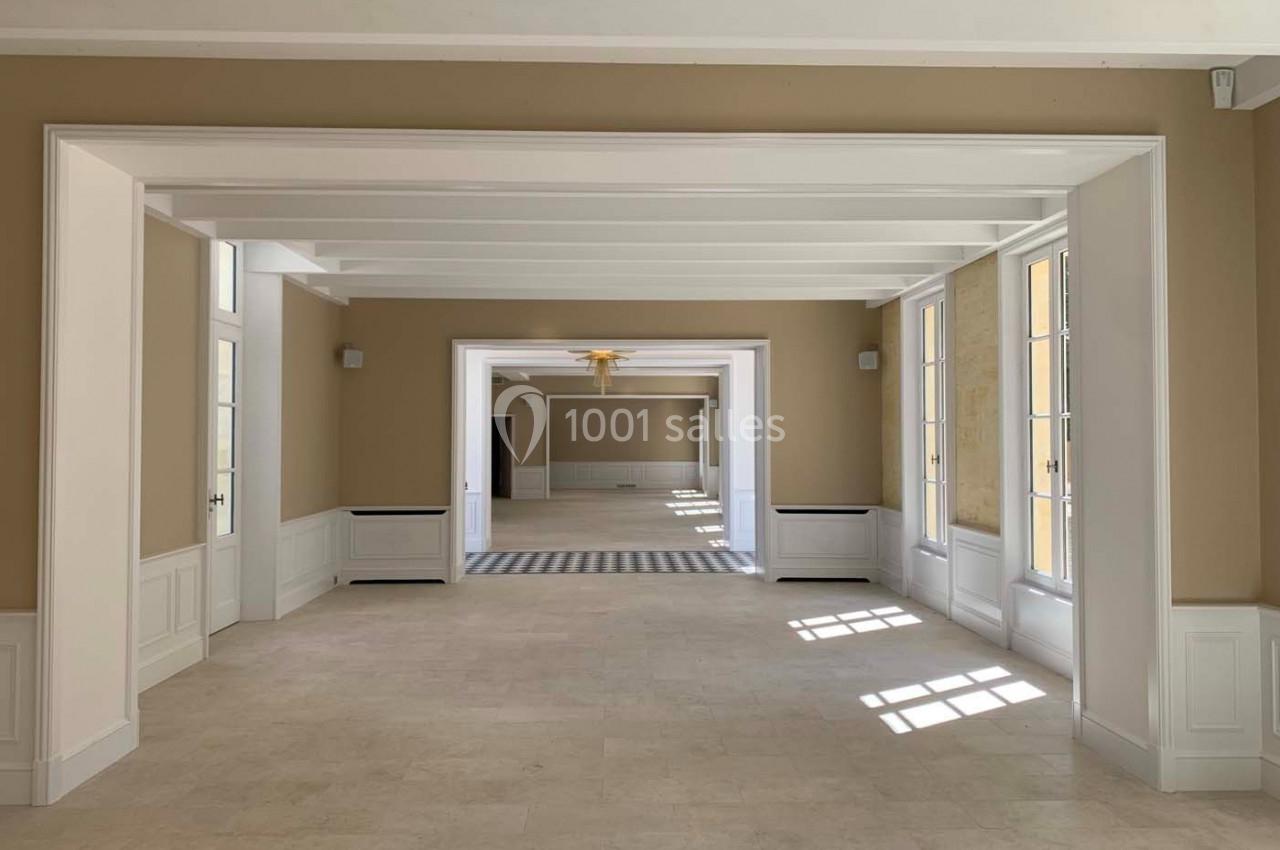 Salle lumineuse avec murs beige, sol en pierre claire, poutres blanches au plafond et grandes fenêtres laissant entrer la…