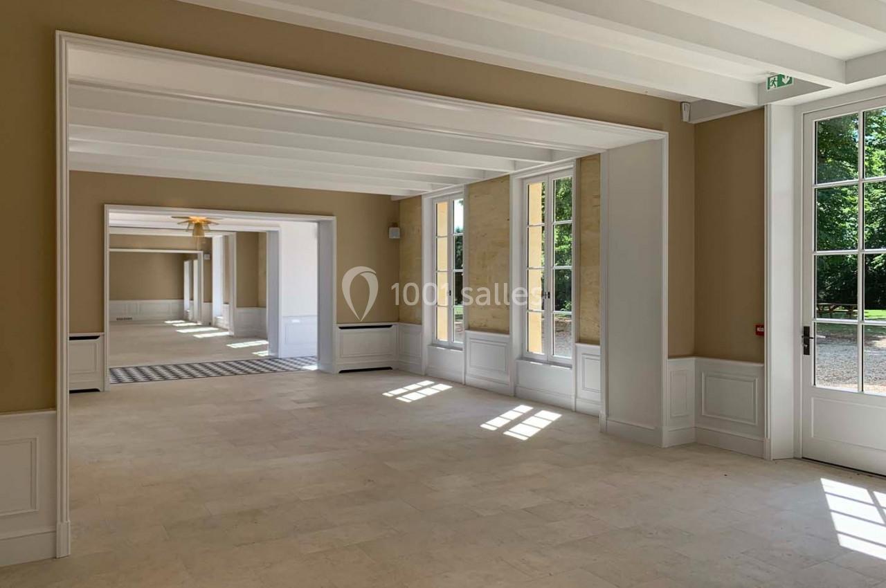 Intérieur lumineux d'une pièce vide avec grandes fenêtres, murs beige clair et sol en pierre, donnant sur un jardin.