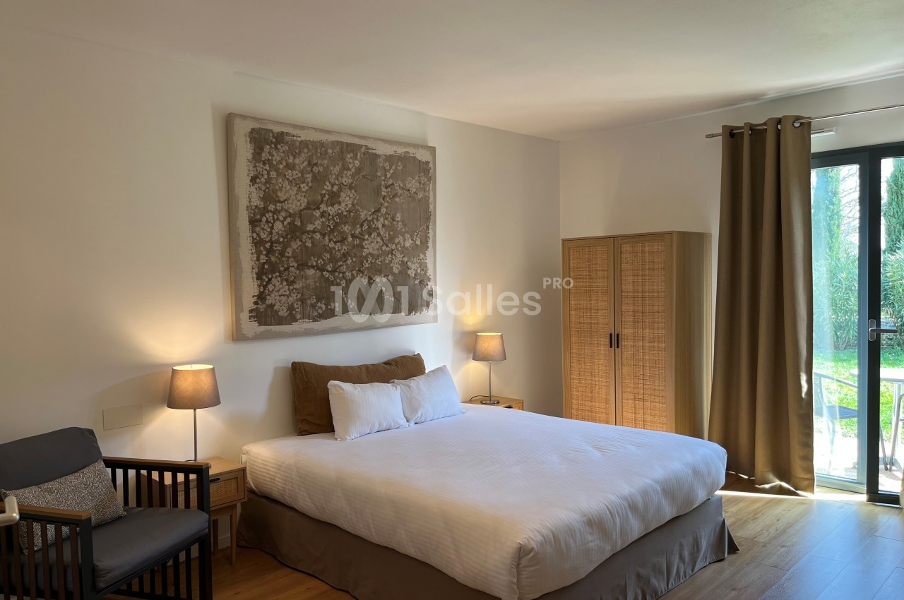 Chambre lumineuse avec lit double, fauteuil, armoire en bois, lampes de chevet et grande fenêtre donnant sur un jardin.