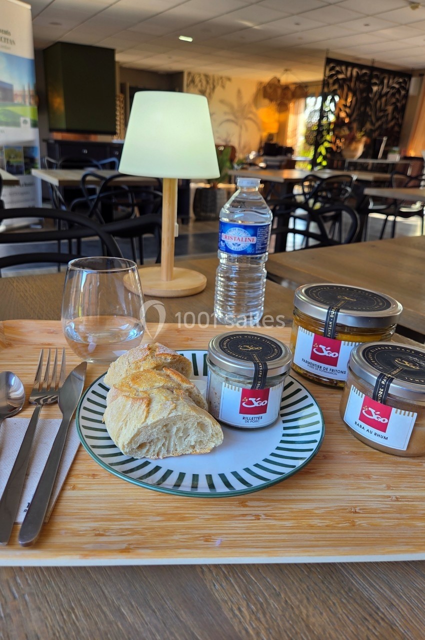 Plateau repas avec pain, pots de confiture, bouteille d'eau, verre vide et couverts sur une table en salle lumineuse.