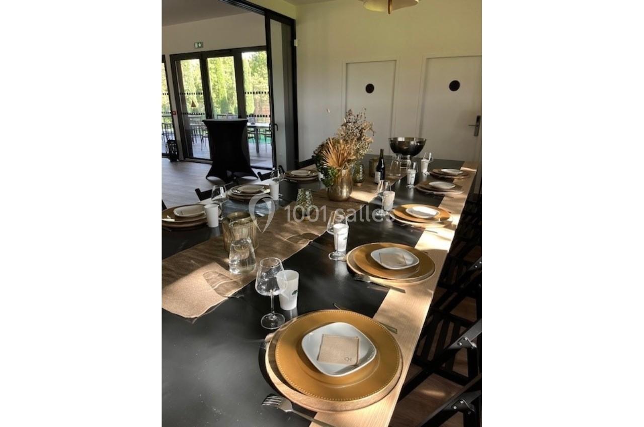 Table dressée avec vaisselle dorée, verres et décoration florale, dans une salle lumineuse avec vue sur l'extérieur.