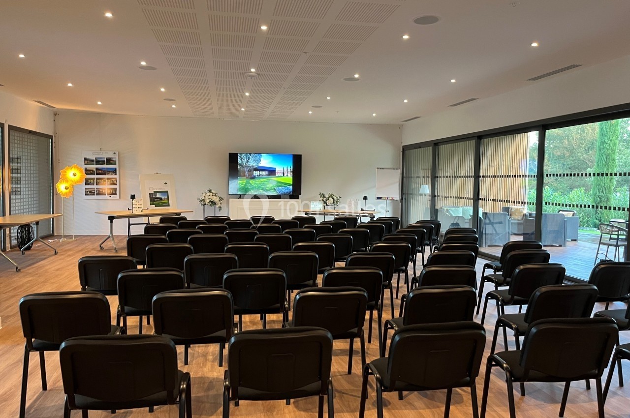 Salle de conférence lumineuse avec des rangées de chaises noires, un écran allumé et des baies vitrées donnant sur un jardin.