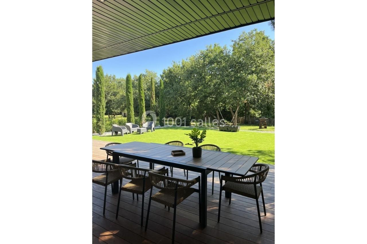 Terrasse en bois avec table et chaises, donnant sur un jardin verdoyant bordé d'arbres et de haies.