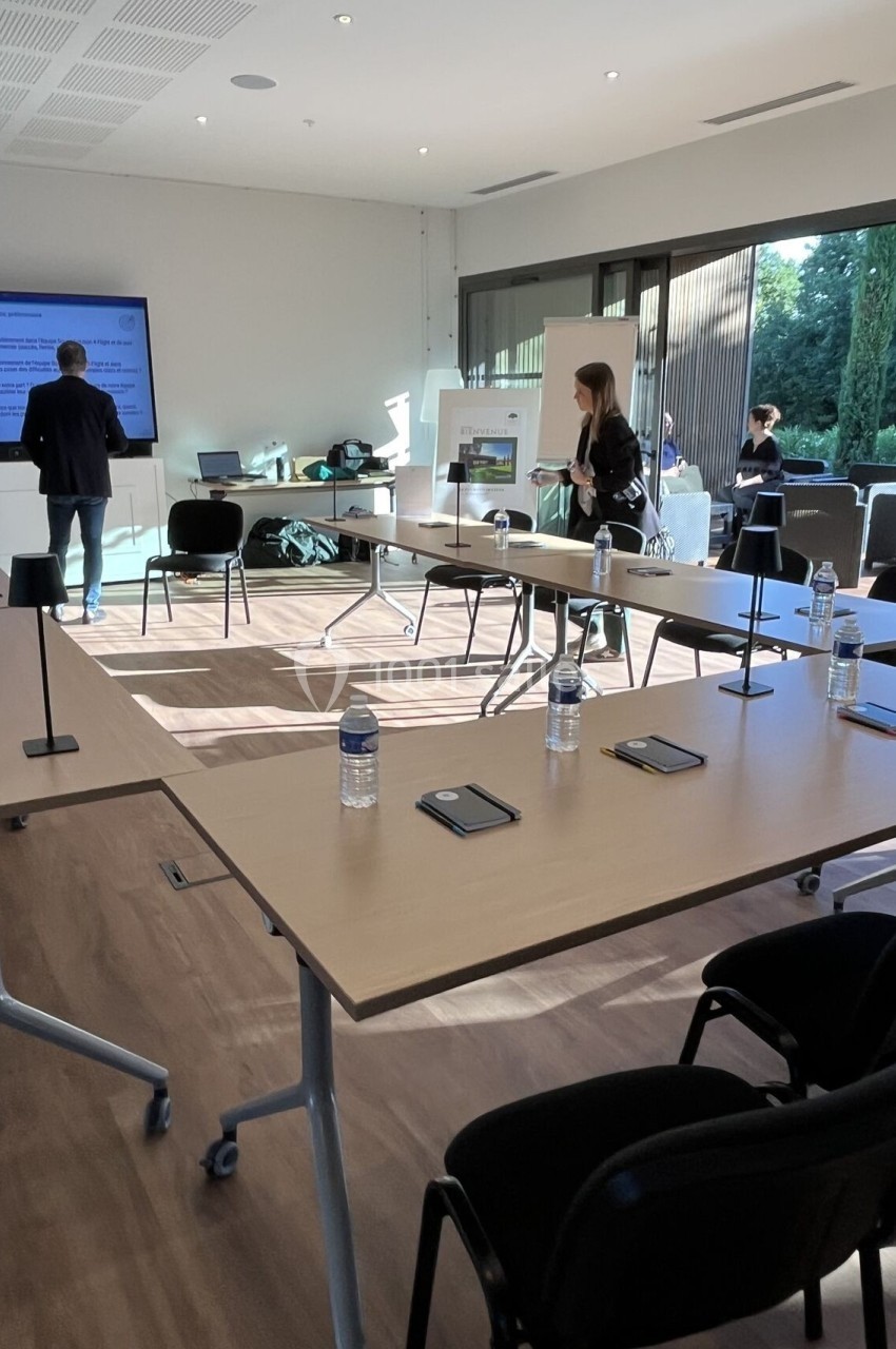 Salle de réunion lumineuse avec tables en U, bouteilles d'eau, écrans et participants en discussion.