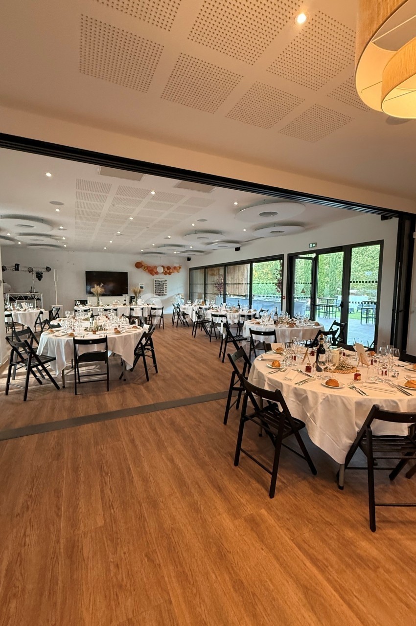 Salle de réception lumineuse avec tables rondes dressées, chaises noires et grandes baies vitrées donnant sur l'extérieur.