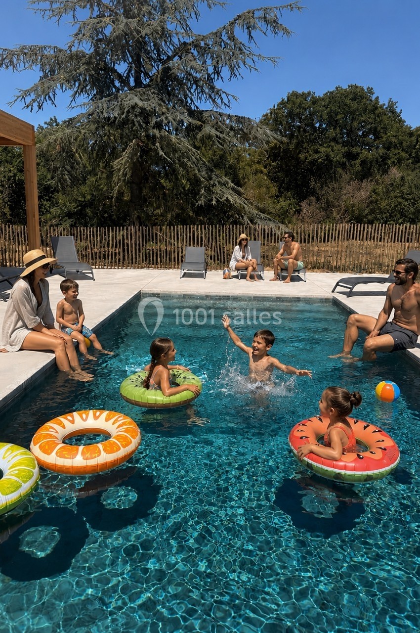 Des enfants jouent avec des bouées dans une piscine, entourés d'adultes assis sur les bords sous un ciel ensoleillé.