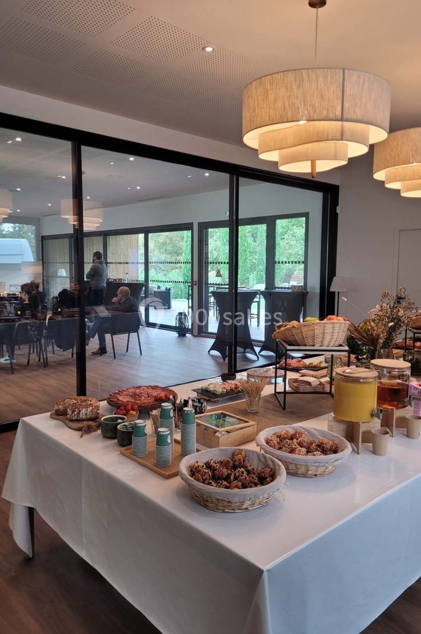 Buffet de petit-déjeuner avec viennoiseries, fruits et boissons, dans une salle lumineuse avec vue sur une terrasse vitrée.