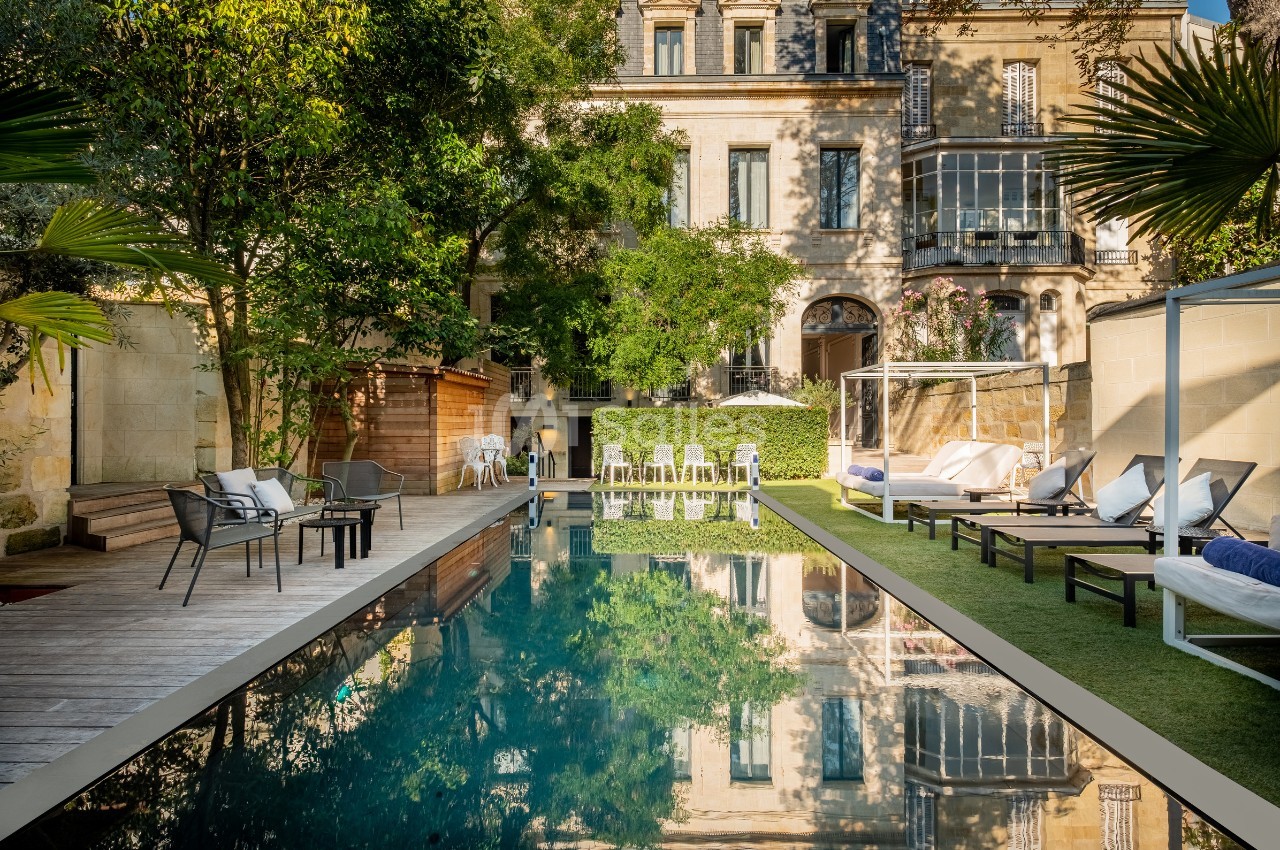 Piscine extérieure entourée de chaises longues, de végétation et d'un bâtiment en pierre avec grandes fenêtres.