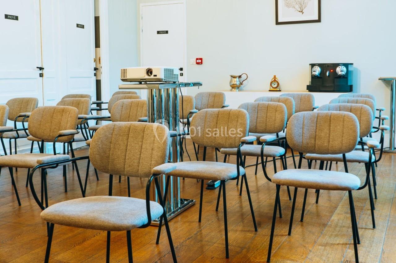 Salle de réunion avec des chaises grises alignées, un projecteur sur un support et un parquet en bois.