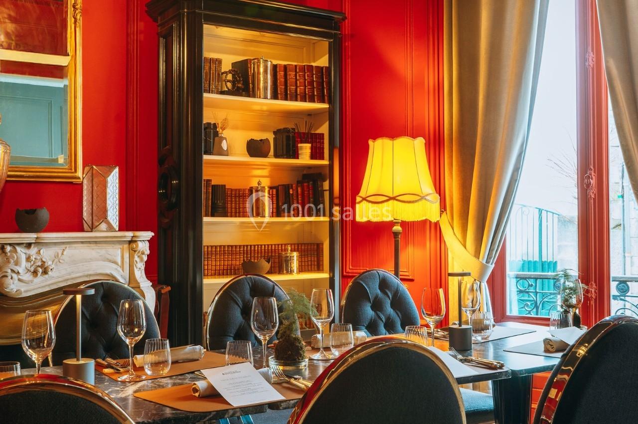 Salle à manger élégante avec murs rouges, bibliothèque éclairée, table dressée et lumière naturelle entrant par la fenêtre.