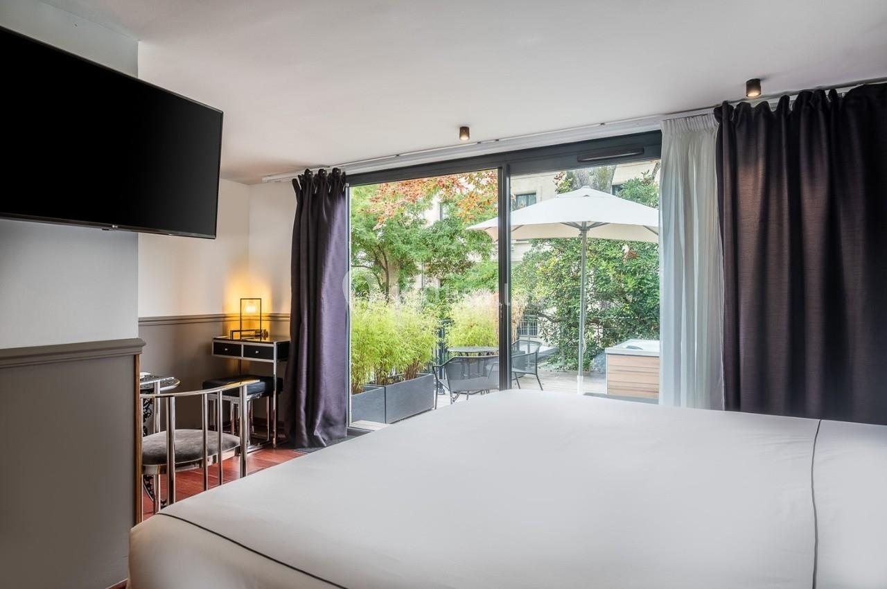 Chambre lumineuse avec lit double, bureau, télévision et accès à une terrasse avec jardin et mobilier extérieur.