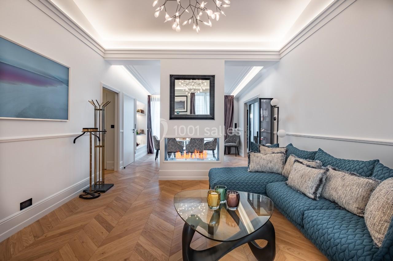 Salon moderne avec parquet en chevrons, canapé bleu, table basse en verre et cheminée intégrée sous un miroir.