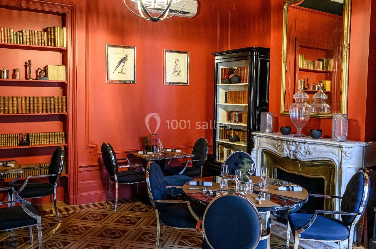 Salle de restaurant élégante avec murs rouges, cheminée ornée, étagères de livres et tables dressées.
