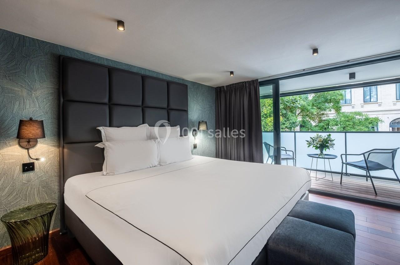 Chambre moderne avec un grand lit, éclairage doux, balcon aménagé et vue sur des arbres à travers une baie vitrée.