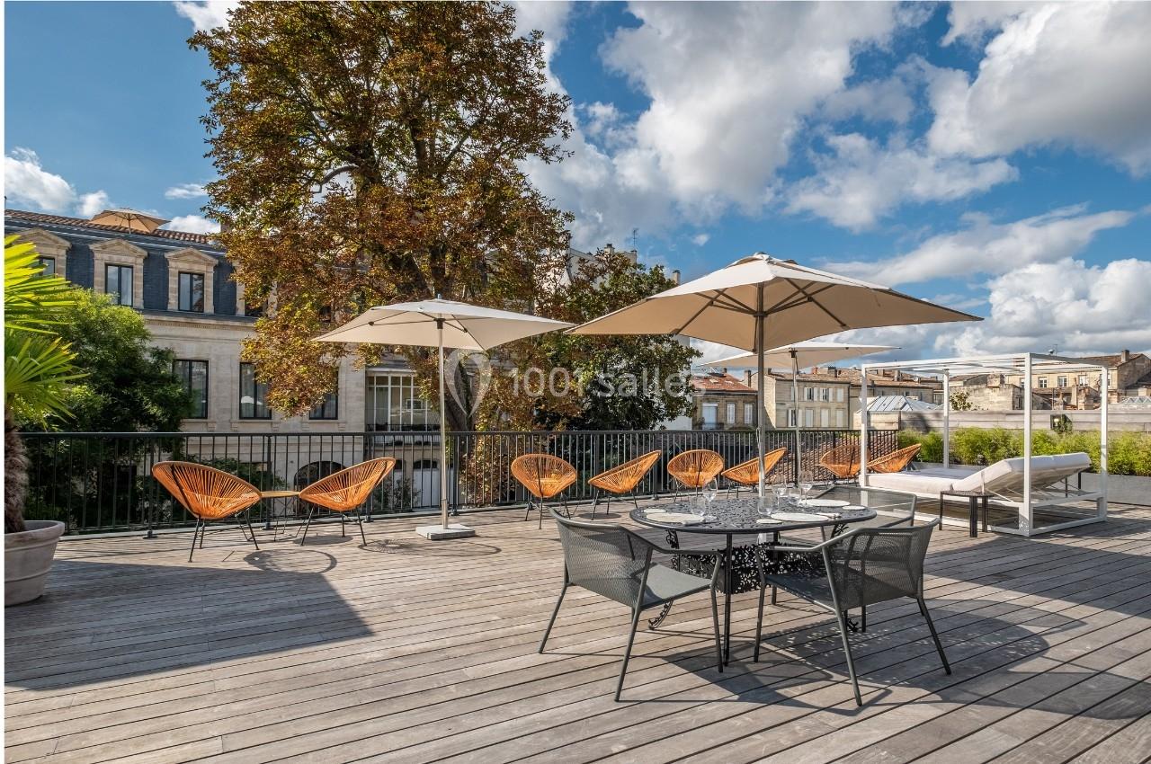 Terrasse en bois avec mobilier de jardin, parasols, chaises et vue sur des bâtiments anciens sous un ciel partiellement…