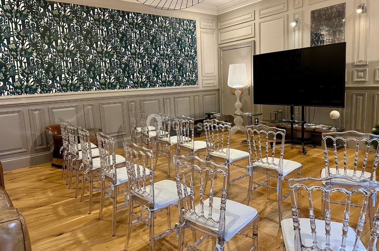Salle avec chaises transparentes alignées, écran noir, lampadaire et mur décoré de boiseries et papier peint végétal.
