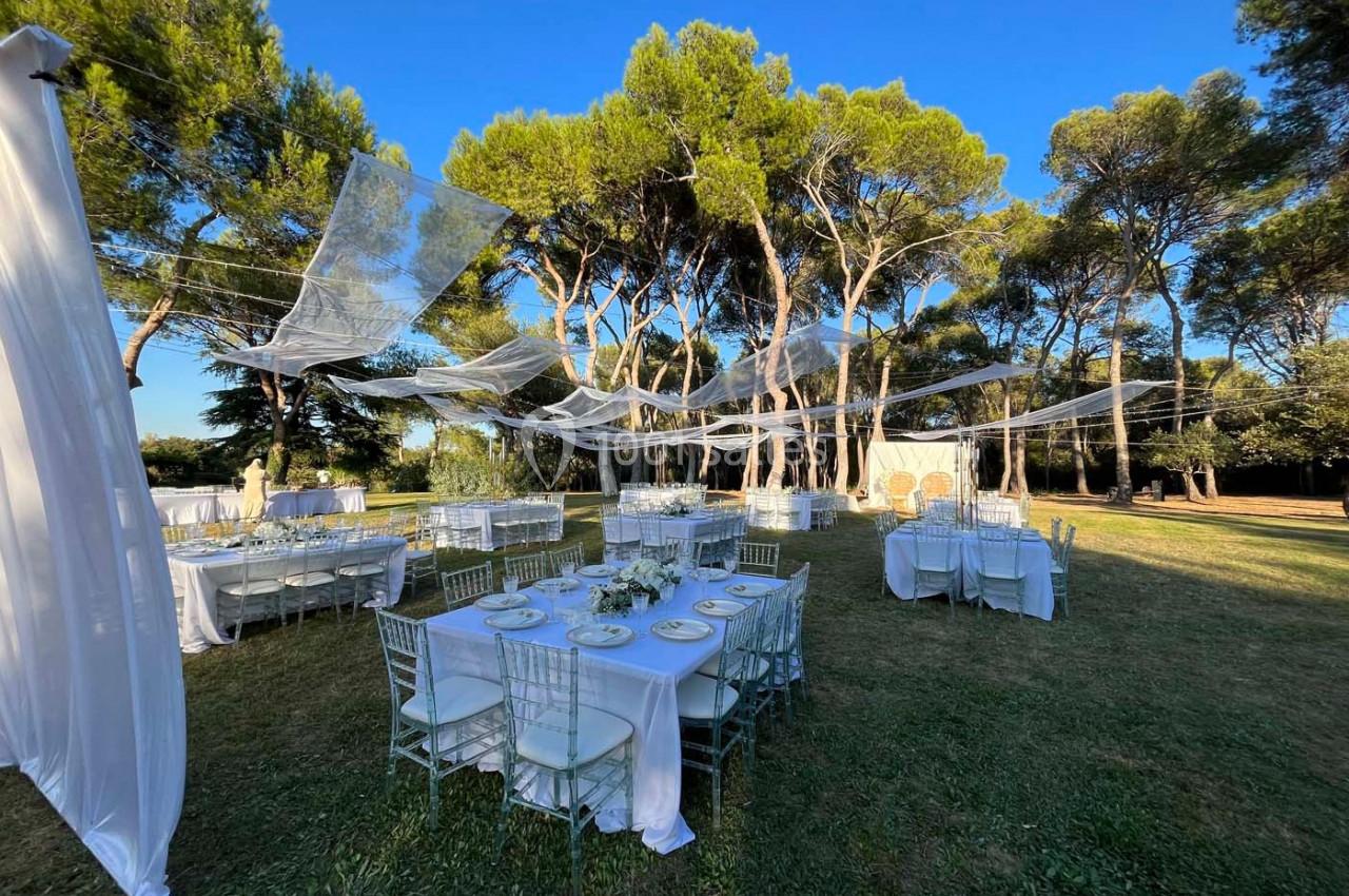 Tables dressées pour un événement en plein air sous des voilages blancs, entourées d'arbres et de pelouse.